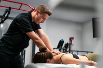 Fisioterapeuta masculino sin afeitar masajeando la espalda de una mujer anónima en la cama durante el procedimiento médico en el hospital - foto de stock