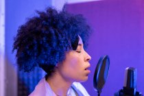 Чорна співачка виконує пісню проти мікрофона з поп-фільтром, стоячи і закриваючи очі в звуковій студії — стокове фото