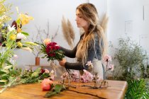 Концентрований молодий жіночий флорист з довгим хвилястим волоссям в повсякденному одязі і фартухах, що влаштовує елегантний букет з троянд і різноманітних квітів в магазині — стокове фото