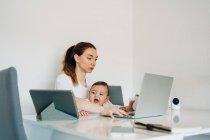 Focused young mother working on laptop holding curious baby watching funny video on tablet while sitting together at desk in light room — Stock Photo