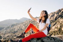 Позитивная молодая женщина-путешественница с кудрявыми темными волосами в повседневной одежде сидит в скалах и улыбается, делая селфи на мобильном телефоне во время похода в горы в солнечный день — стоковое фото