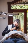 Kosmetikerin mit Pinzette beim Auftragen gefälschter Wimpern zur Verlängerung auf das Auge eines ethnischen Kunden mit Gesichtsschutzmaske im Salon während der Coronavirus-Pandemie — Stockfoto