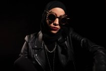 Впевнена молода мусульманка в шкіряній куртці, хустка і творчі сонцезахисні окуляри сидять на чорному тлі в студії — стокове фото