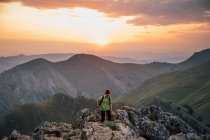Weite Wanderin auf hohem Felsgipfel gegen majestätische Bergkette unter wolkenverhangenem Himmel im Sonnenuntergang — Stockfoto