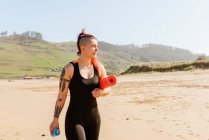 Atleta sonriente con alfombra enrollada y botella de agua paseando por la costa arenosa del mar mientras mira hacia otro lado - foto de stock