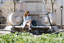 Полное тело молодая африканская американка в синем пальто просматривает на мобильном телефоне, сидя на каменной скамейке в весеннем парке возле скутера — стоковое фото