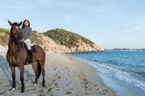 Молода жінка дивиться далеко під час їзди на каштановому коні на піщаному морському березі проти гори під небом — стокове фото