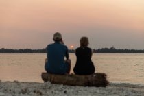 Rückansicht Unerkennbares Paar sitzt auf Baumstamm am sandigen Meeresufer und genießt malerischen Sonnenuntergang über ruhigem plätscherndem Meer — Stockfoto