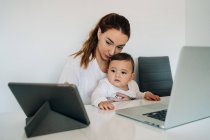 Орієнтована молода мати працює на ноутбуці, тримаючи допитливу дитину, дивлячись смішне відео на планшеті, сидячи разом за столом у світлій кімнаті — стокове фото