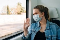 Giovane donna che indossa maschera medica e saluto amico in stazione mentre cavalca in treno e sorride — Foto stock