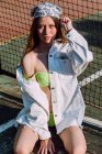 Giovane turista femminile sensuale in costume da bagno e bandana toccare i capelli mentre guardando la fotocamera contro recinzione griglia — Foto stock
