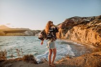 Back view anonymous fit female in mini skirt standing on rough rocky seashore and touching long hair under clear blue sky in Fyriplaka Milos — Stock Photo