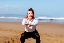 Весела жінка-спортсменка тренується з простягнутими руками на піщаному узбережжі океану, дивлячись на камеру — стокове фото