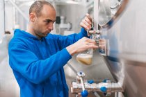 Seitenansicht eines männlichen Arbeiters in steriler Maske, der in der Fabrik fermentiertes Bier aus einem Metallgefäß in Glas gießt — Stockfoto
