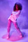 Comprimento total apto jovem dançarina afro-americana em desgaste informal solto olhando para a câmera enquanto dança no estúdio escuro em luzes de néon — Fotografia de Stock