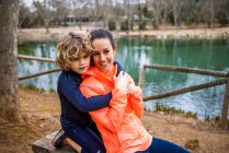 Charming boy embracing smiling mother while sitting and looking away against water in daylight — Foto stock