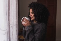 Вид сбоку очаровательной этнической женщины с черными афро-волосами, стоящей с чашкой напитка дома и выглядывающей в окно — стоковое фото
