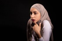 Modish fiducioso femminile musulmana in hijab appoggiato sulla sedia e guardando altrove in studio scuro — Foto stock