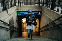 Ethnic female in hijab and protective mask walking upstairs in city street while using smartphone — Stock Photo