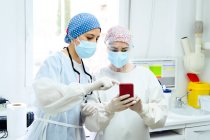 Cirurgiões do sexo feminino em tampas de pano médico navegar na internet no celular contra computador desktop no hospital luz — Fotografia de Stock