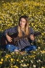 Задумчивая молодая хипстерша, сидящая на лугу в сельской местности и играющая на гитаре в летний солнечный день — стоковое фото