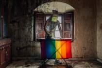 Unrecognizable person in silver costume and box on head ironing LGBTQ flag while standing in shabby abandoned room — Stock Photo