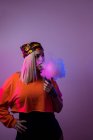 Cool hembra en traje de calle fumar cigarrillo electrónico y exhalación de humo a través de la nariz y la boca en el fondo púrpura en el estudio con iluminación de neón rosa - foto de stock