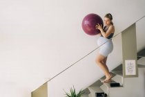 Вид збоку на молоду усміхнену майбутню жінку в спортивному одязі з м'ячем йоги, що прогулюється по сходах в будинку — стокове фото