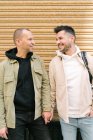 Positive young multiethnic gays in stylish clothes smiling and looking at each other while holding hands on city street — Foto stock