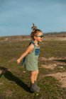 Vista lateral do conteúdo menina em macacões e óculos de sol pulando com os braços estendidos acima do prado e desfrutando de verão no dia ensolarado no campo — Fotografia de Stock