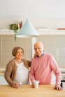 Content middle aged couple standing with cups of drinks in kitchen while hugging and looking at camera — Stock Photo