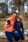 Cheerful mom in sports clothes embracing charming boy while taking self portrait on cellphone in park — Foto stock