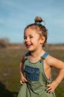 Encantada adorável menina em macacão de pé com as mãos na cintura no prado e olhando para longe — Fotografia de Stock