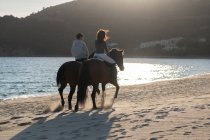 Back view of unrecognizable young woman with boyfriend riding purebred stallions on sandy shore against wavy ocean under blue sky — Stock Photo
