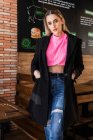 Attraente giovane donna in jeans maglione rosa e cappotto nero in piedi con le mani in tasche vicino al tavolo nel ristorante e guardando la fotocamera — Foto stock