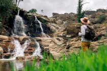 Back view anonymous male backpacker in hat enjoying view of cascade streaming from rough rock in verdant nature — Stock Photo
