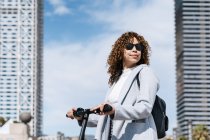 Optimistic young African American female with curly hair wearing blue coat and sunglasses standing with scooter on city street on sunny spring weather — Stock Photo