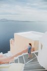Von oben steht eine gesichtslose junge Frau in eleganter Kleidung auf alten Steintreppen in einem authentischen Küstenort mit weißen Häusern und bewundert das plätschernde blaue Meer in Griechenland — Stockfoto