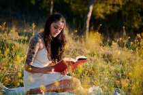 Dreamy charming brunette in white dress sitting on field meadow and reading book in sunlight — Foto stock
