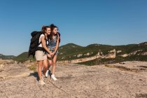 Delighted female hikers with backpacks cuddling on rock in highlands and looking away while enjoying summer vacation — Stock Photo