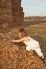 Вид сбоку симпатичной маленькой девочки в комбинезоне, лазающей по соломенному тюку во время игры вечером в сельской местности — стоковое фото