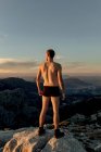 Voltar ver anônimo caminhante masculino em cuecas pretas em pé no cume da montanha rochosa e admirando paisagens montanhas espetaculares ao pôr do sol — Fotografia de Stock