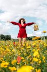 Dal basso felice femmina in abito da sole rosso, cappello e borsetta in piedi con gli occhi chiusi su campo fiorito con fiori gialli e rossi con le braccia tese godendo nella calda giornata primaverile estiva — Foto stock
