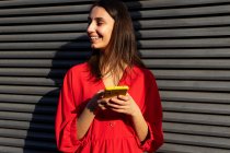 Молодий контент жінки в червоному вбранні балакає на мобільному телефоні на сонячному світлі на сірому фоні — стокове фото