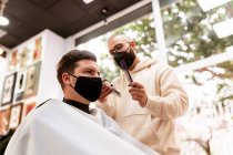 Männlicher Friseur mit Brille schneidet erwachsenen Kunden im Friseursalon während der COVID-19-Pandemie die Haare — Stockfoto