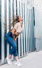 Side view of millennial female with tattoos and long Afro braids leaning forward against wall on urban walkway — Stock Photo