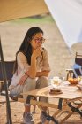 Bella femmina asiatica etnica in occhiali da sole seduta a tavola a bere tè mentre si gode un momento di relax in campeggio durante le vacanze — Foto stock