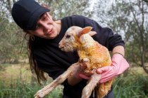 Agricultora alegre em desgaste preto e luvas carregando pequeno cordeiro recém-nascido no quintal de verão — Fotografia de Stock