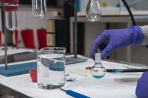 Анонимный ученый в защитных перчатках, трясущий синим химическим веществом во фляжке во время работы в современной оборудованной лаборатории — стоковое фото
