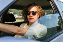 Serious young haired male in stylish sunglasses looking at camera through open window of car while sitting at driver seat — Stock Photo